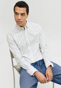 Jeune homme aux cheveux courts et bouclés, portant une chemise blanche à rayures et un jean bleu, assis sur une chaise, regardant sur le côté.