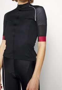 Maillot de cyclisme noir ajusté avec manches courtes à motifs mesh et poignets rouges, assorti à un cuissard de cyclisme noir rembourré.