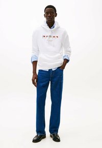 Witte hoodie met kleurrijk logo, blauwe overhemd eronder, gecombineerd met blauwe jeans en zwarte instappers. Eenvoudig ontwerp en relaxte pasvorm.