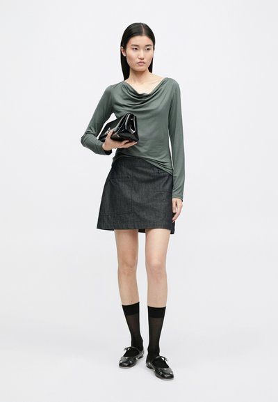 Blusa verde oliva de mangas longas com decote em cowl, combinada com uma saia escura de ganga. Clutch preta brilhante, meias pretas até ao joelho e mocassins.