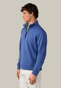 Jeune homme face à gauche porte un pull bleu roi à fermeture éclair quart de zip avec un logo sur la manche, une chemise vert clair en dessous et un pantalon blanc.