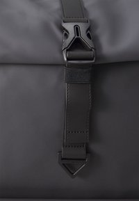 Sac à fermeture roulante noir avec une large sangle et une grande boucle triangulaire en plastique.