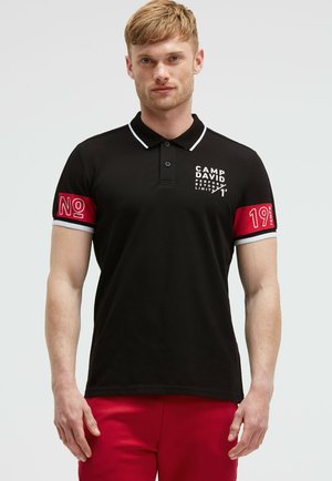 Zwarte polo shirt met witte kraag, voorzien van een geprint logo op de borst en rode accenten op de mouwen met de tekst "Nee" en "19".
