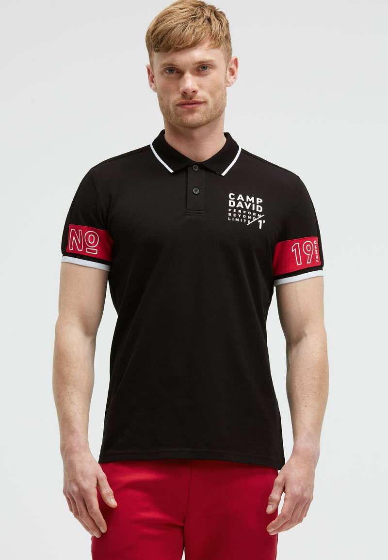 Polo nero con colletto bianco, caratterizzato da un logo stampato sul petto e dettagli rossi sulle maniche con il testo "No" e "19".
