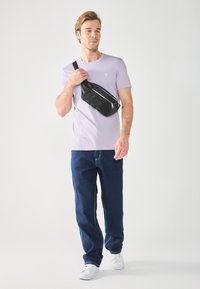 Black fabric waist bag with silver zipper details, worn over a light purple t-shirt and dark blue jeans. White sneakers complete the look.