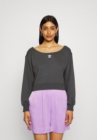 Sweatshirt cropped de cor cinza escuro com um decote largo e mangas longas, apresentando o logótipo branco da Adidas. Combinado com calções soltos de cor roxa clara.