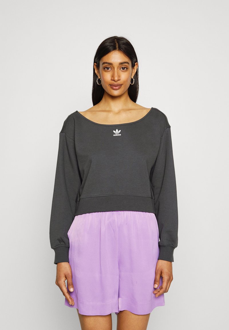 Sweatshirt cropped de cor cinza escuro com um decote largo e mangas longas, apresentando o logótipo branco da Adidas. Combinado com calções soltos de cor roxa clara.