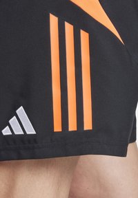 Zwarte sportshorts met drie verticale oranje strepen en een klein zilveren driehoeklogo op de linkerbenedenkant. Glad stoffen textuur.