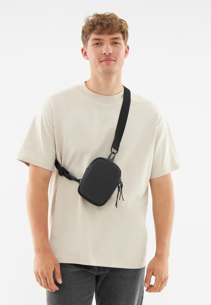 Borsa a tracolla nera realizzata in materiale liscio, di forma rettangolare, con tracolla regolabile e scomparti con zip. Indossata sopra una camicia beige.