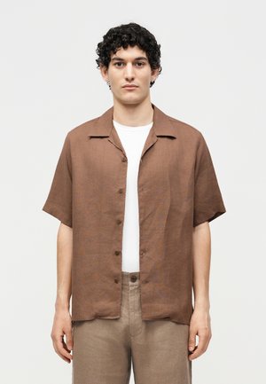 Jeune homme aux cheveux courts et bouclés portant une chemise marron à manches courtes par-dessus un t-shirt blanc et un pantalon beige, debout devant un fond uni.