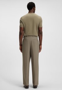 Man wearing olive green short-sleeve shirt tucked into matching tailored trousers, standing facing away on a plain white background.