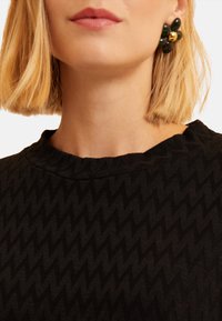 Prenda negra texturizada con un patrón de zigzag y un escote redondo, combinada con pendientes florales en verde y dorado.