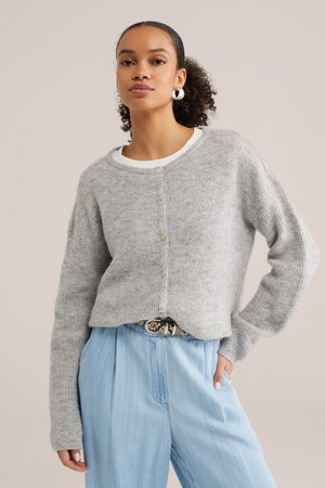 Ženska, oblečena v svetlo sivi gumbasti pulover, belo majico, svetlo modre hlače z visokim pasom in obročaste uhane, pozira z eno roko v žepu.