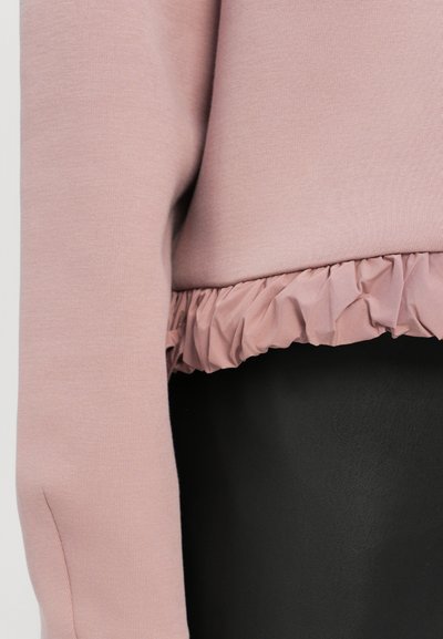 Heled roosa sviiter, millel on tekstuuriga volangserv, pikad varrukad ja siledat materjali. Kanga välimus on pehme ja selle stiil on juhuslik.