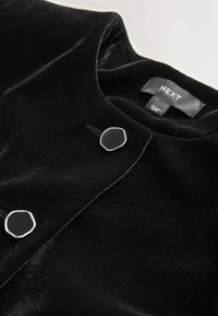 Veste en velours noire avec un col rond, deux boutons hexagonaux noirs aux bords argentés et une étiquette de marque « NEXT » à l’intérieur du col.