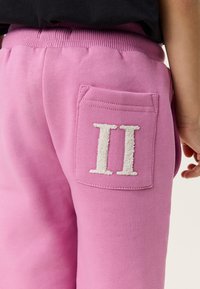 Κοντινό πλάνο σε ροζ παντελόνι με πίσω τσέπη που διαθέτει λευκό υφασμάτινο λατινικό αριθμό II και μέρος του χεριού ενός ατόμου.