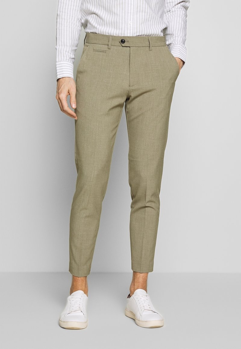 Homme portant un pantalon slim marron clair, une chemise blanche rayée et des baskets blanches, debout avec une main dans la poche.