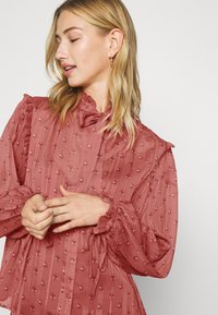 Blusa ligera y con volantes en rosa empolvado, con patrones florales texturizados y un lazo en el cuello; presenta mangas largas con puños con volantes.
