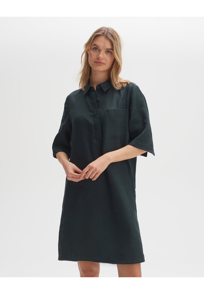 Donkergroen linnen shirtjurk met een knoopsluiting bij de kraag, korte mouwen en een enkele borstzak. Soepele textuur en een ontspannen pasvorm.