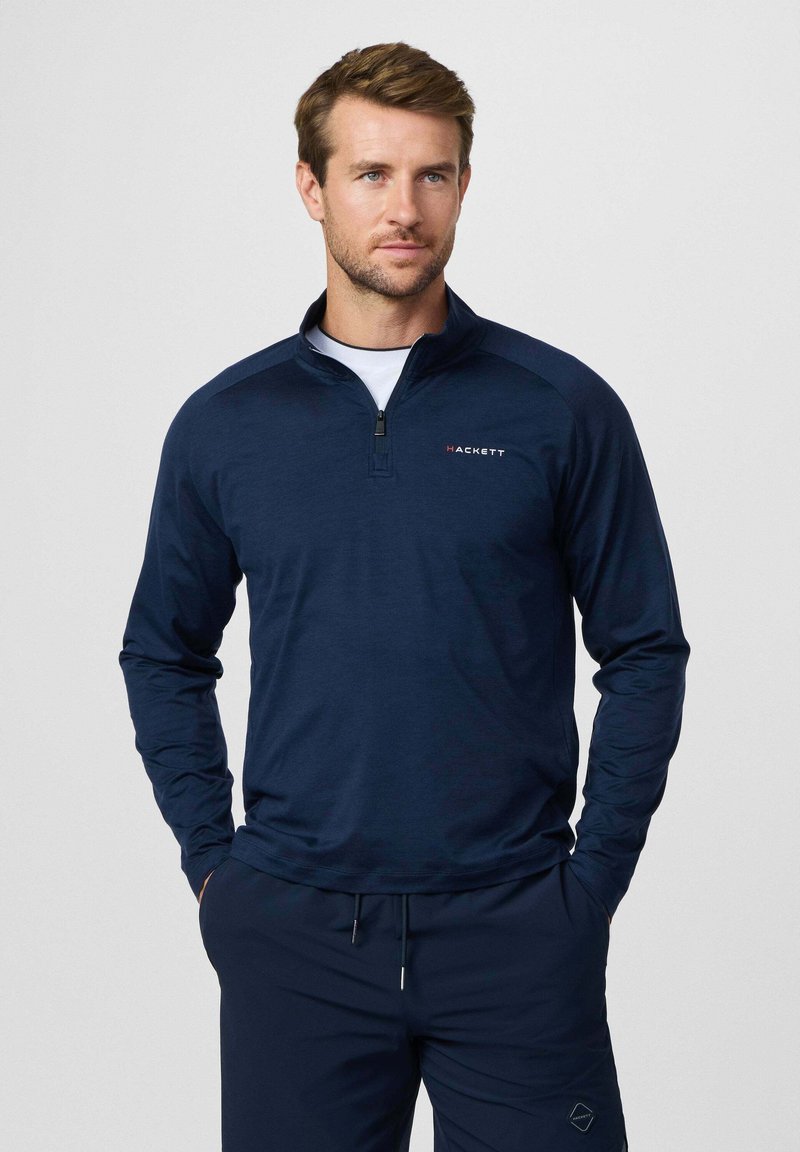 Uomo che indossa un pullover blu navy con zip a quarto e logo "Hackett" e pantaloni blu coordinati, in piedi con le mani nelle tasche contro uno sfondo semplice.