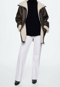 Giacca in ecopelle marrone con foderatura in shearling crema, dolcevita nera in maglia, pantaloni bianchi a gamba larga e scarpe nere con punta affusolata.