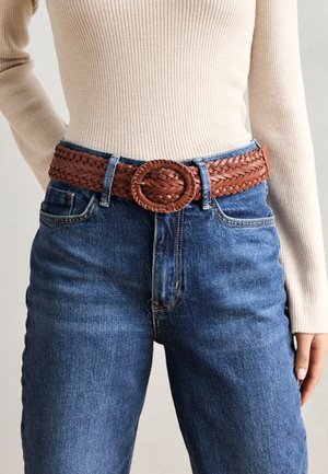 Curea din piele împletită, de culoare maro, cu o cataramă rotundă, purtată peste blugi din denim albastru, cu talie înaltă, având cusături minime și un design clasic.