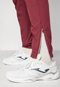 Sarkanas šauras bikses ar rāvējslēdzēja galiem, apvienotas ar baltām sporta kurpēm. Biksēm ir gluda virsma un elastīga josta.