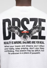 Aigle noir aux ailes déployées au-dessus des grandes lettres "DRSZE" et texte motivant au dos d'un t-shirt blanc porté par une personne.