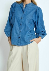 Džínová košile na knoflíky světle modré barvy s límečkem do stoje, nabranými rukávy a náprsní kapsou. Kombinováno s krémovými širokými kalhotami.