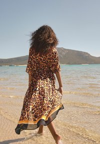 Maksi haljina sa leopard uzorkom od lepršavog, prozirnog materijala, s kratkim rukavima i crno-žutim rubom, koju nosi figura na plaži.