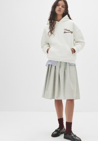 Felpa bianca con testo marrone "Più di un'etichetta", indossata sopra una camicia a righe azzurre. Gonna grigia a pieghe, calze borgogna e scarpe nere.