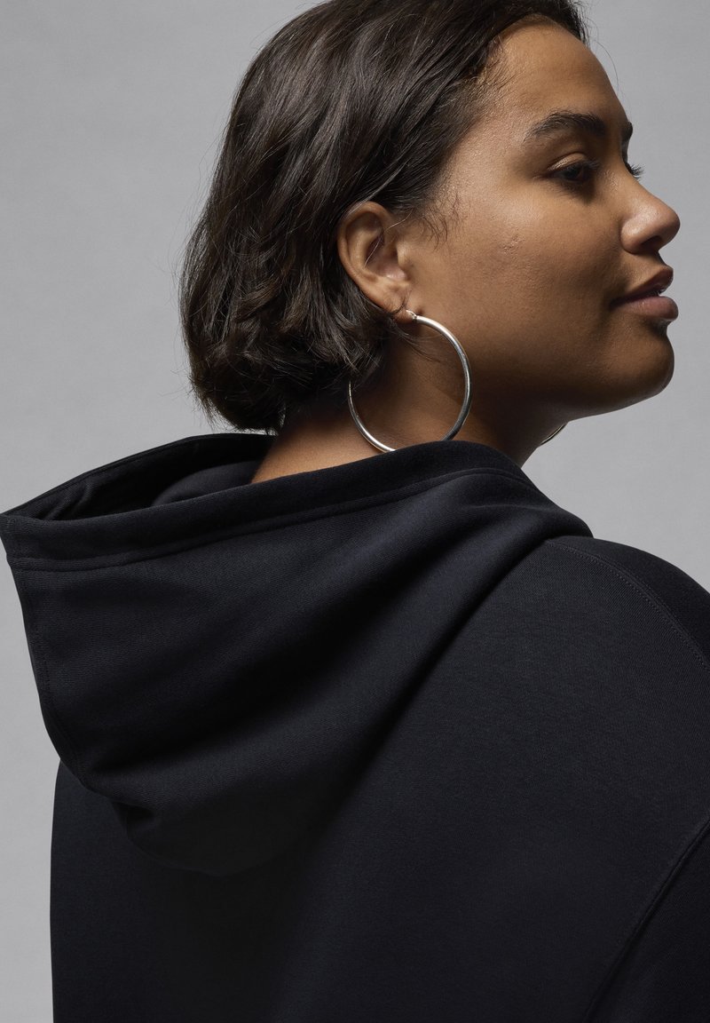 Haut à capuche noir avec cordon de serrage et tissu texturé. Le modèle porte de grandes boucles d'oreilles en argent, mettant en valeur un gros plan de son profil latéral.
