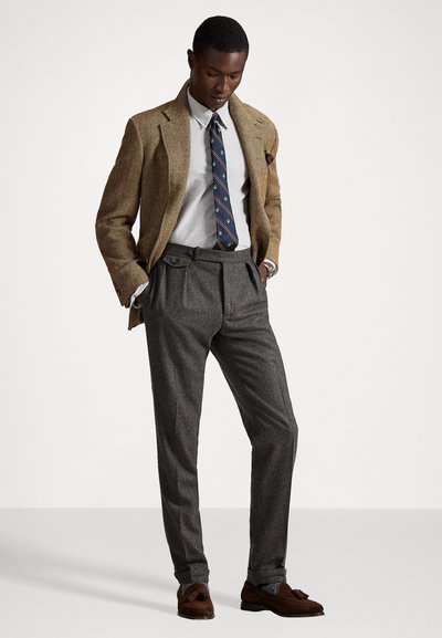 Brūns herringbone žakets, balts svītrainā krekls, zils rakstains kaklasiksna, pelēkas plīša bikses, brūni mokasīni. Pielāgota forma ar elegantiem akcentiem.