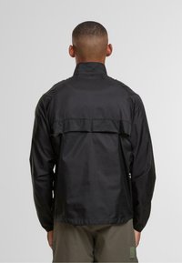 Uomo con i capelli corti, indossante una giacca a vento nera e pantaloni verde oliva, visto da dietro su uno sfondo grigio uniforme.