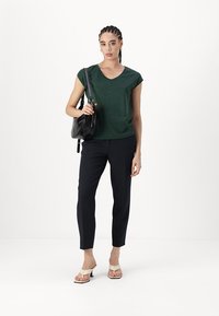 Dark green short-sleeve top, navy tailored pants, and off-white strappy heels. Model carries a black shoulder bag. Minimalist style.