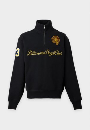 Jeune homme portant un sweat noir à fermeture quart de zip avec un texte jaune et un patch, un jean bleu à jambes larges, et des chaussures noires et blanches, debout avec une main dans la poche.