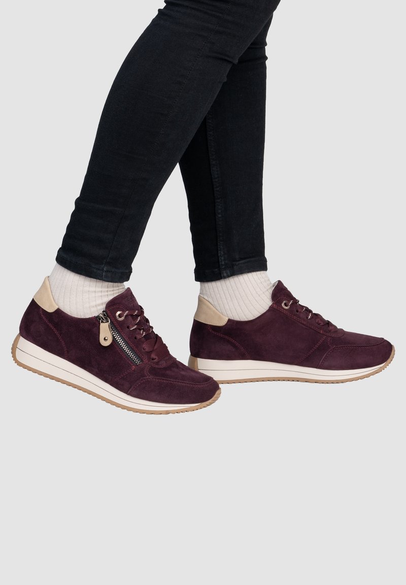 Personne portant des baskets en daim bordeaux avec des accents beige et des chaussettes blanches côtelées, associées à un pantalon noir ajusté.