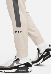 Joggers de color beige claro con una franja gris oscura, con "AIR" impreso en negro. Combinados con zapatillas Nike Air Max en blanco y negro.