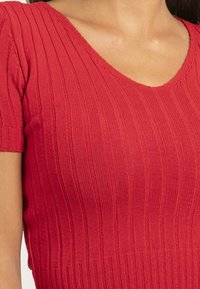 Haut en tricot côtelé rouge à manches courtes, doté d'un décolleté en V et d'une texture douce, mettant en valeur les lignes verticales et l'élasticité.
