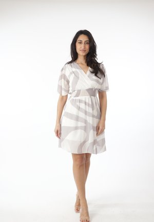 Femme debout, portant une robe blanche longueur genoux avec des motifs abstraits gris clair et des sandales à talons transparents sur un fond blanc.