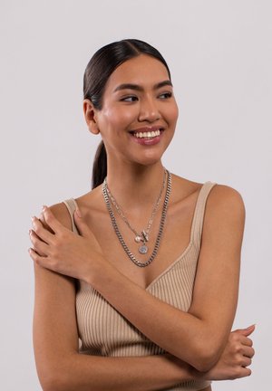Zilveren gelaagde halskettingen met ketting- en bedeldetails, gedragen door een model met lang, donker haar, in een beige geribbelde top tegen een neutrale achtergrond.