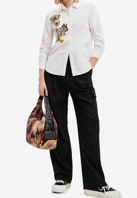 Bílá košile s knoflíky a květinovou výšivkou, černé kalhoty s širokými nohavicemi a vzorovaná kabelka s černým popruhem a drobnými cvočky.