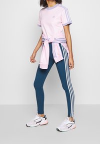 Ljusrosa t-shirt med lavendelaccenter, knuten i midjan, ihop med teal leggings med vita sidosträck och vita sneakers med svarta detaljer.