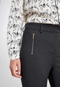 Pantalon noir avec une poche zippée, associé à une chemise blanche ornée d'un motif de cygne noir, présentant une coupe ajustée et une texture lisse.