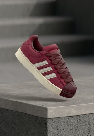 Bordowy zamszowy sneaker z gumowym noskiem, z trzema białymi paskami, grubymi sznurowadłami oraz teksturowaną śródstopiem na szarej powierzchni.