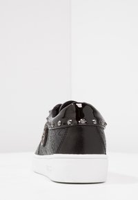 Sneaker nera con suola bianca, collo del tallone lucido con borchie e motivo in rilievo, mostrata da dietro su uno sfondo bianco.