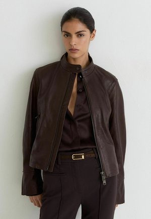 Donna con capelli scuri che indossa una giacca di pelle marrone scuro sopra una camicia scura e pantaloni scuri, in piedi contro una parete chiara.