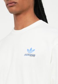 Artimas kadras asmens, kuris dėvi baltą Adidas Originals marškinėlį su mėlynu trefoil logotipu ir tekstu ant krūtinės.