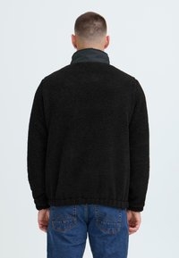 Mann iført svart strukturert fleecejakke og blå jeans, vist fra ryggen mot en enkel lys bakgrunn.
