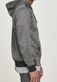 Personne portant un sweat à capuche gris texturé avec des poignets et une ceinture noirs, debout de profil, les mains détendues le long du corps.
